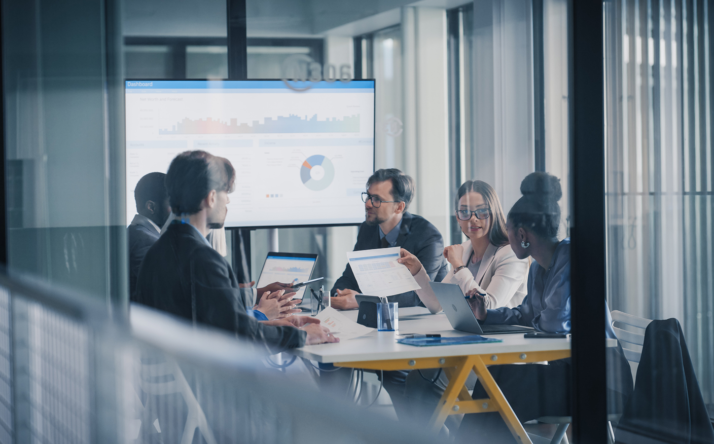 Six people sit around a conference table with laptops and documents, discussing data shown on a screen with charts and graphs in a modern office setting.