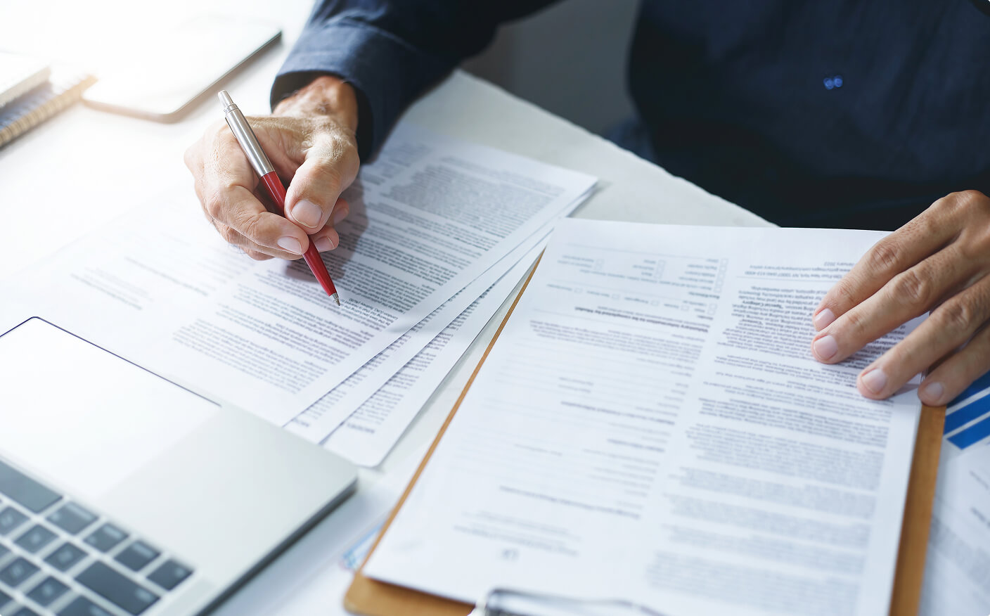 A person reviews and fills out paperwork at a desk with a laptop, clipboard, and pen, appearing focused on the documents in front of them.